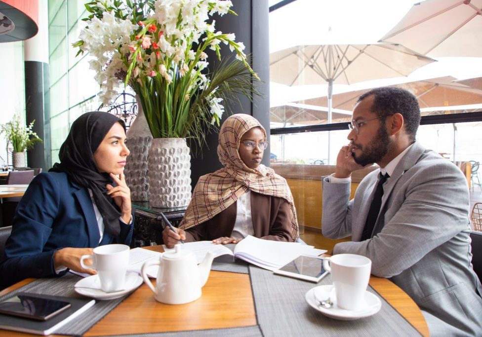 Multiethnic business partners working on agreement in cafe. Businessman and Muslim businesswomen sitting in coffee shop, calling on phone and signing documents. Partnership concept